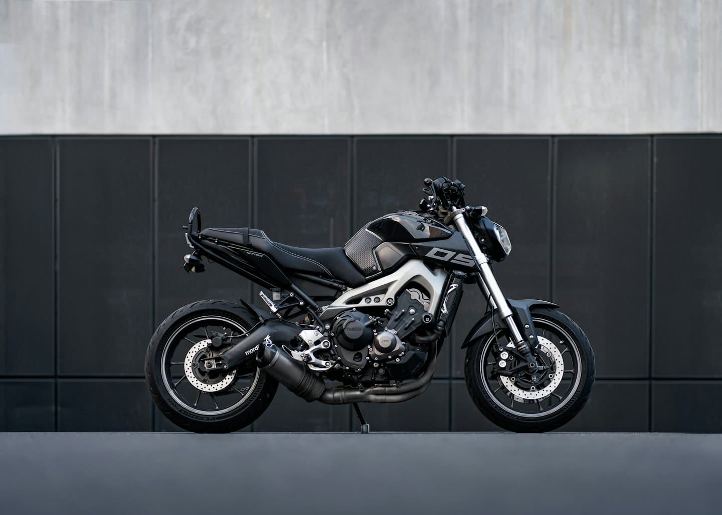 Black motorcycle parked against a minimalist grey concrete wall.